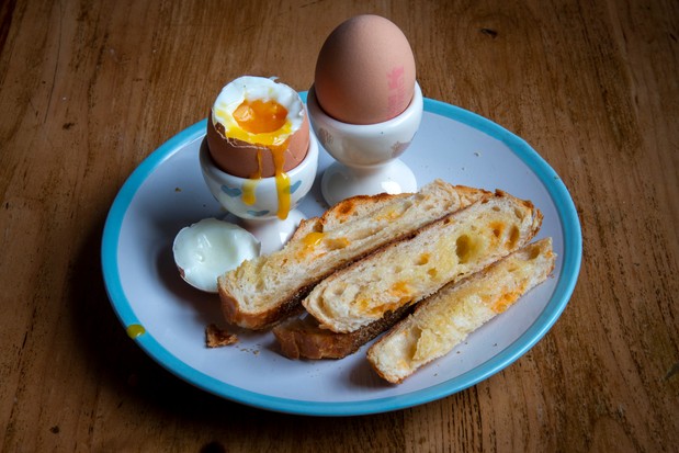 Deux œufs durs dans des coquetiers avec un jaune qui coule et trois soldats en pain de fromage grillé sur une assiette blanche à bords bleus sur une table en bois.