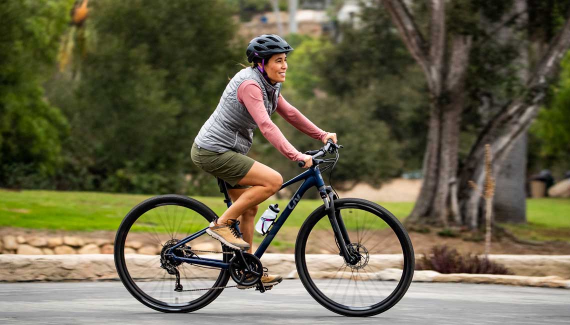 Une femme de couleur roule sur un vélo hybride, souriante, le long de la route. Elle porte un short, un gilet et un casque.