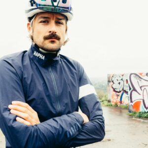 A white man with his arms crossed in a blue waterproof cycling jacket looks directly into the camera