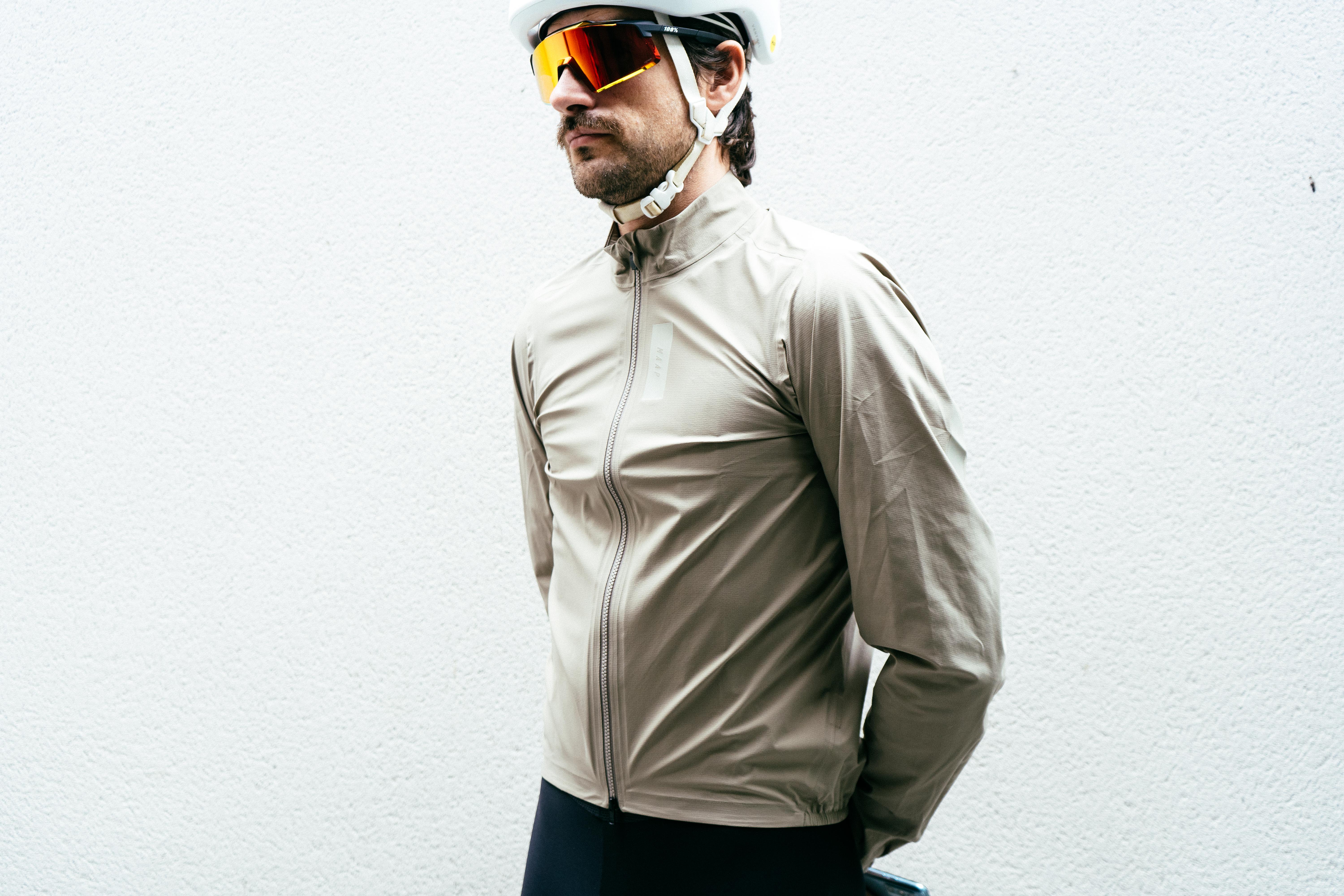 Un homme vêtu d'une veste de pluie blanc cassé, d'un casque blanc et de lunettes de soleil orange contre un mur blanc