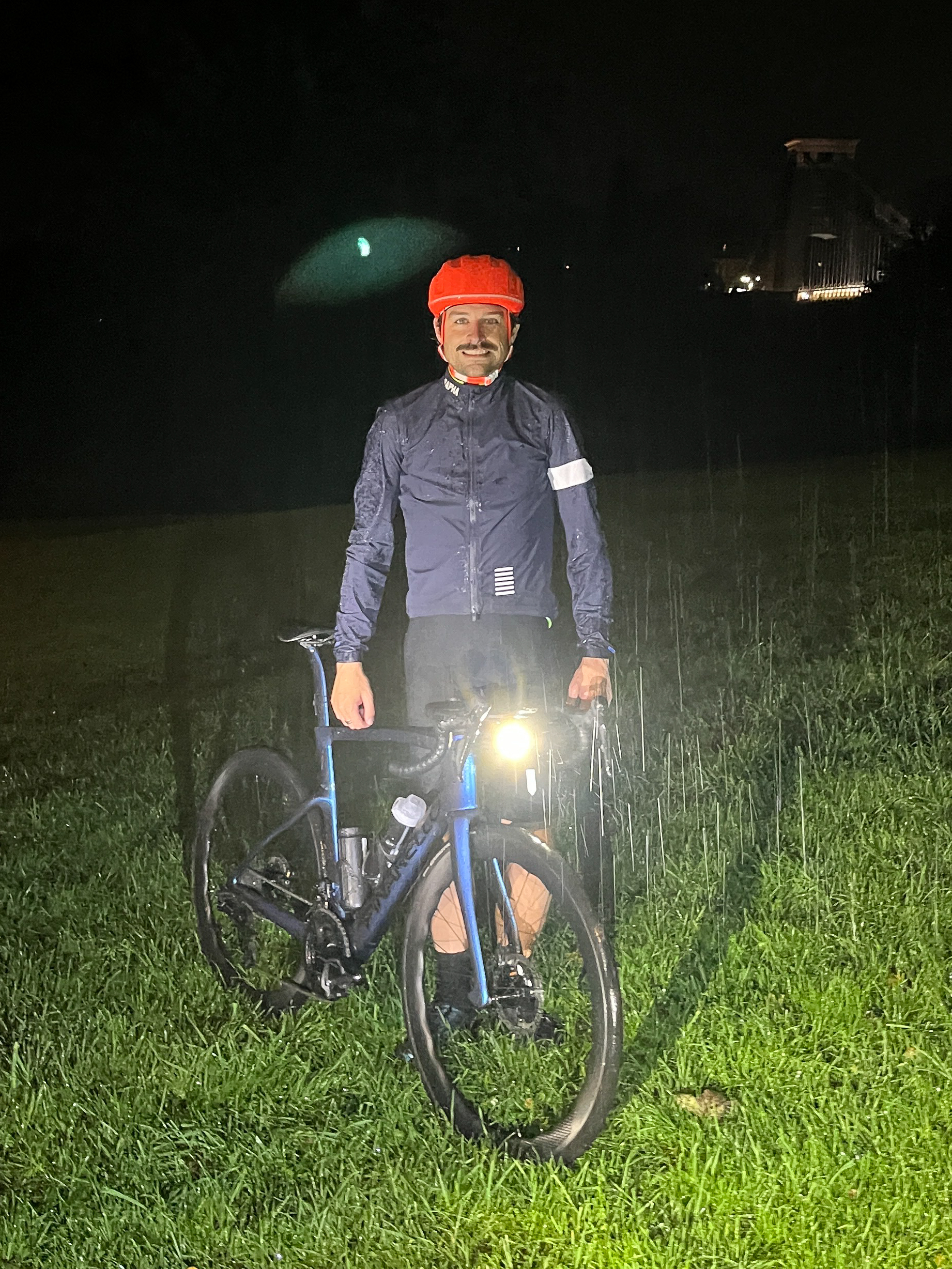 Un homme blanc vêtu d'une veste de pluie bleue et d'un casque orange se tient sous la pluie à côté d'un vélo