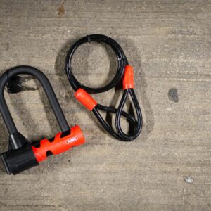 A black and red Abus Ultimate 420 lock and cable on a stone floor next to a bike rack