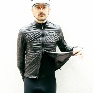 A white man wears a thin balck insulated cycling jacket against a white wall