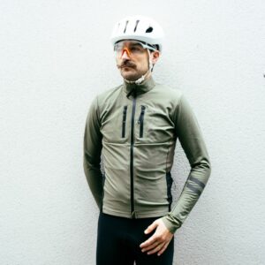 A white man in an olive green winter cycling jacket stands against a grey wall